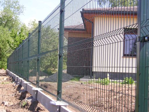 Zielone ogrodzenie panelowe z podmurówką betonową na nierównym terenie, widok z boku na tle domu z beżową elewacją i zielenią.