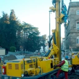 Żółta wiertnica na gąsienicach, obsługiwana przez pracownika w kasku i kamizelce odblaskowej, na tle budynku z kamienia i drzew.