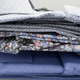 Stos starannie złożonych męskich koszul z różnymi wzorami: drobna kratka, motyw kwiatowy i regularne geometryczne desenie w odcieniach bieli i błękitu.