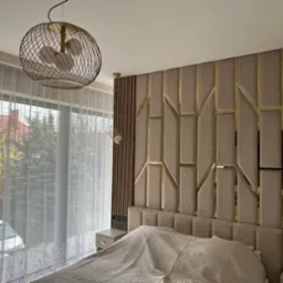 Sypialnia z dekoracyjnym panelem ściennym w beżowych odcieniach z geometrycznymi złotymi akcentami, oświetlona wiszącymi lampami z metalową konstrukcją, widok na okno z białymi zasłonami i drzewami...