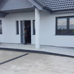 Ułożona kostka betonowa przed wejściem do domu z białymi ścianami i szarą dachówką, widoczny pracownik w drzwiach.
