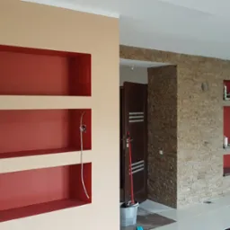 Ściana z wnękami pomalowanymi na czerwono, jedna część ściany w kolorze beżowym, druga wyłożona kamieniem dekoracyjnym, widoczne przewody elektryczne i akcesoria malarskie.