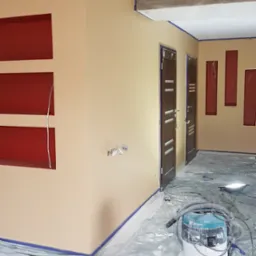Pokój w trakcie malowania: ściany pomalowane na beżowo, z wyciętymi wnękami pomalowanymi na czerwono, zabezpieczone folią i taśmą malarską, widoczny odkurzacz przemysłowy i wiadro z farbą.