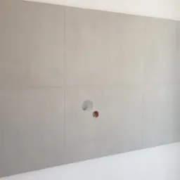 Ściana łazienki wyłożona szarymi płytami imitującymi beton z otworem na instalację, w rogu wnęka z półkami.