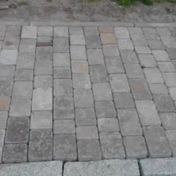 Szara kostka ułożona w regularny wzór, obrzeże z większych kamieni, widoczny fragment młotka brukarskiego w tle.