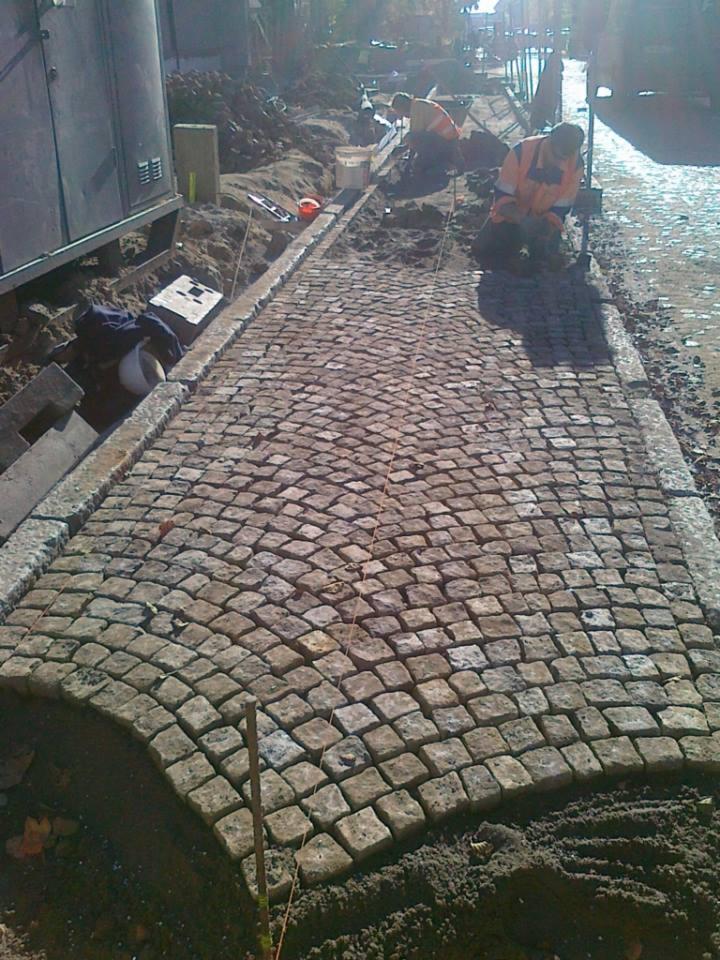 Fragment nowo układanej drogi z kostki granitowej w kształcie wachlarza, widoczni robotnicy w pomarańczowych kamizelkach oraz sprzęt budowlany w tle.