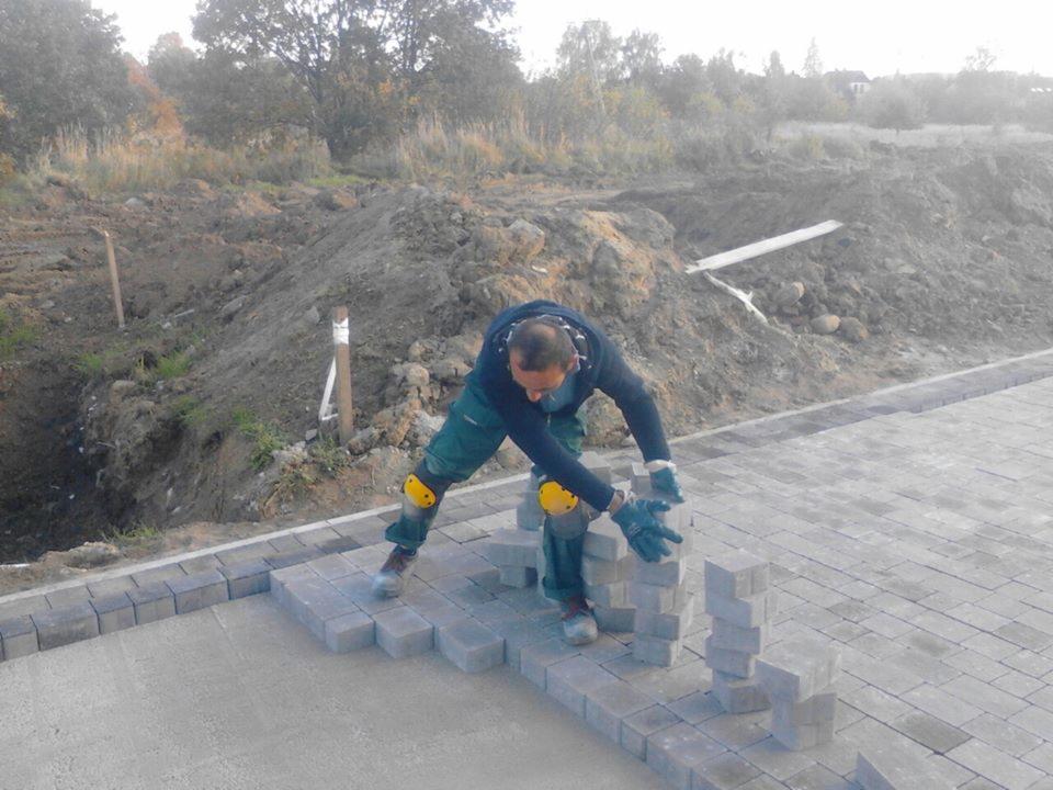 Pracownik w stroju roboczym z żółtymi nakolannikami układa szare kostki brukowe na przygotowanym podłożu, w tle widoczne wykopane ziemie i drzewa.