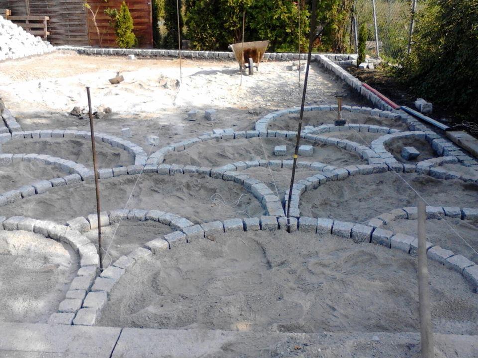 Etap formowania wzoru z kostki granitowej w piasku, widoczne sznurki wyznaczające kształt, wbite pręty i częściowo ułożone obrzeża.