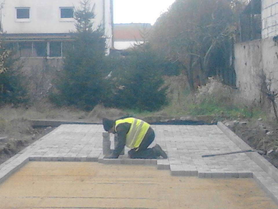 Pracownik w żółtej kamizelce układa szarą kostkę brukową na przygotowanym podłożu z piasku, widoczne obrzeża i budynek mieszkalny w tle.