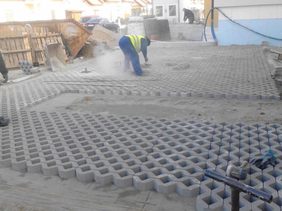 Montaż ażurowej kostki brukowej na podsypce piaskowej, widoczny pracownik w odblaskowej kamizelce, gumowy młotek i rękawice robocze, w tle kontener na gruz.