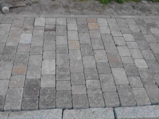 Szara kostka ułożona w regularny wzór, obrzeże z większych kamieni, widoczny fragment młotka brukarskiego w tle.