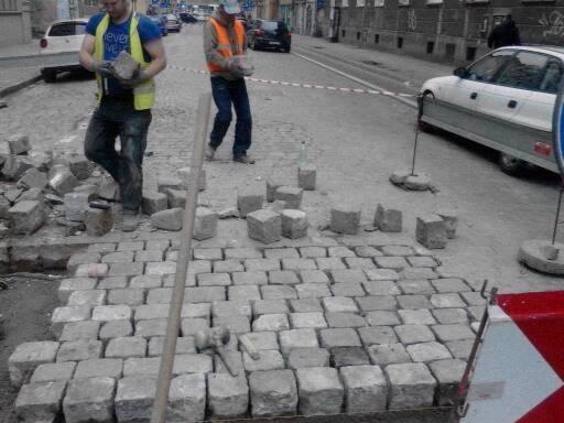 Dwóch robotników układa kostkę granitową na ulicy, widoczne narzędzia i częściowo ułożona nawierzchnia, zabezpieczona taśmą ostrzegawczą, w tle zaparkowane samochody i budynki miejskie.