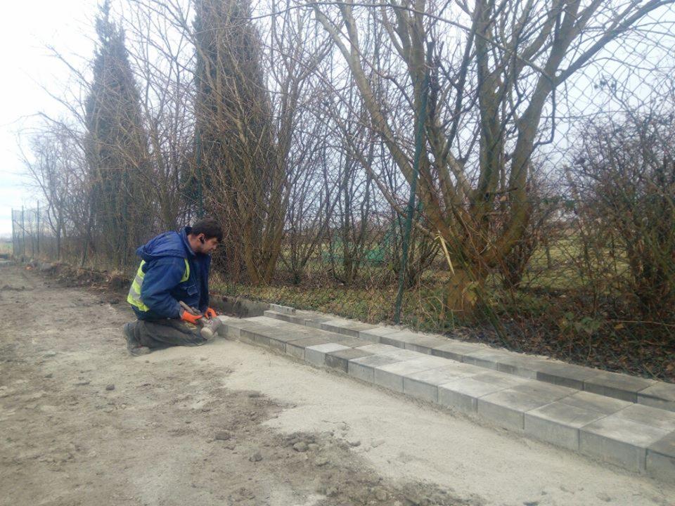 Mężczyzna w odblaskowej kamizelce klęczy i wbija gumowym młotkiem szarą kostkę brukową w podsypkę piaskową, tworząc ścieżkę wzdłuż ogrodzenia.