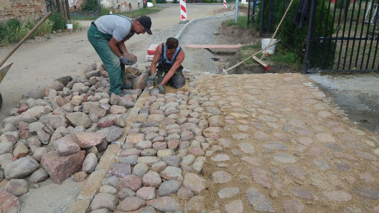 Dwóch mężczyzn w trakcie układania nawierzchni z nieregularnych kamieni polnych, część już ułożona i zasypana piaskiem, widoczny fragment drogi w tle.