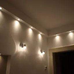 Nowoczesne oświetlenie sufitowe z punktowymi lampami LED i dwoma kinkietami ściennymi z elementami kryształowymi w jasnym pomieszczeniu. Widoczny fragment ściany z gładzią oraz górna część futryny...