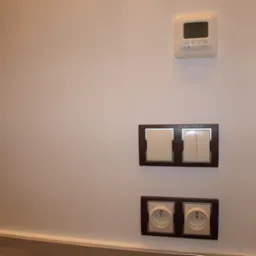 Ściana z gładzi pomalowana na biało z zamontowanym termostatem, podwójnym włącznikiem światła i gniazdkami elektrycznymi w ciemnobrązowych ramkach.