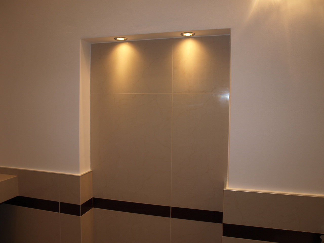 Wnęka w łazience wyłożona beżowymi płytkami, oświetlona dwoma punktowymi lampami halogenowymi, z widocznym fragmentem dolnej części ściany z ciemnym pasem dekoracyjnym.