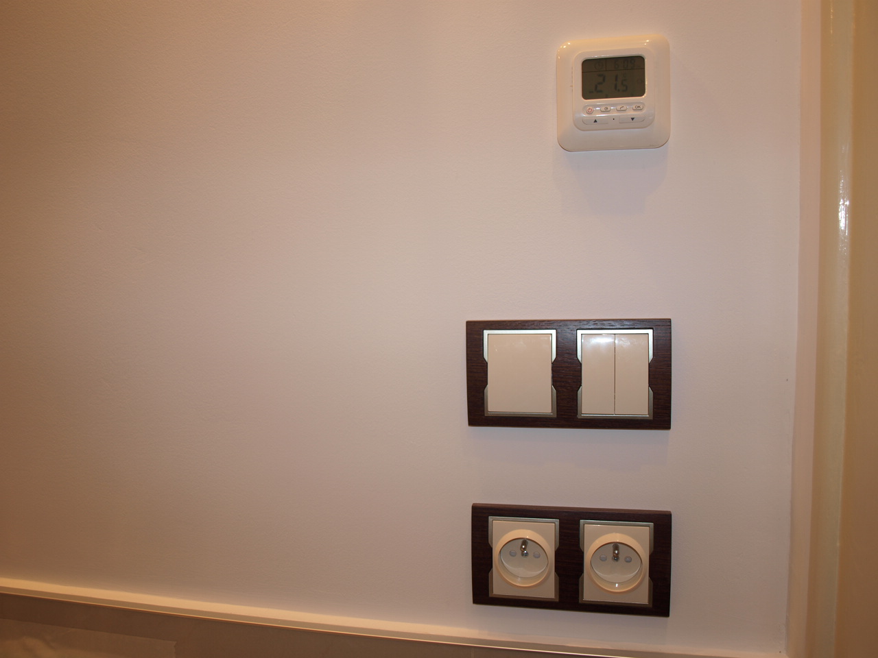 Ściana z gładzi pomalowana na biało z zamontowanym termostatem, podwójnym włącznikiem światła i gniazdkami elektrycznymi w ciemnobrązowych ramkach.