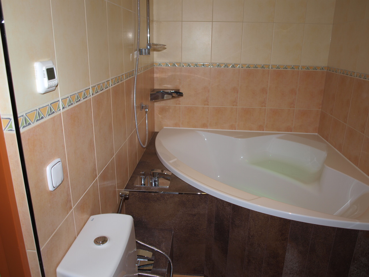 Jasna łazienka z beżowymi i brązowymi płytkami, narożną wanną z hydromasażem wypełnioną wodą, oraz wiszącą toaletą z chromowaną słuchawką bidetową.