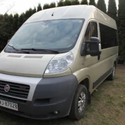 Beżowy bus Fiat Ducato z ciemnymi szybami, zaparkowany na trawiastym terenie, z widoczną tablicą rejestracyjną.