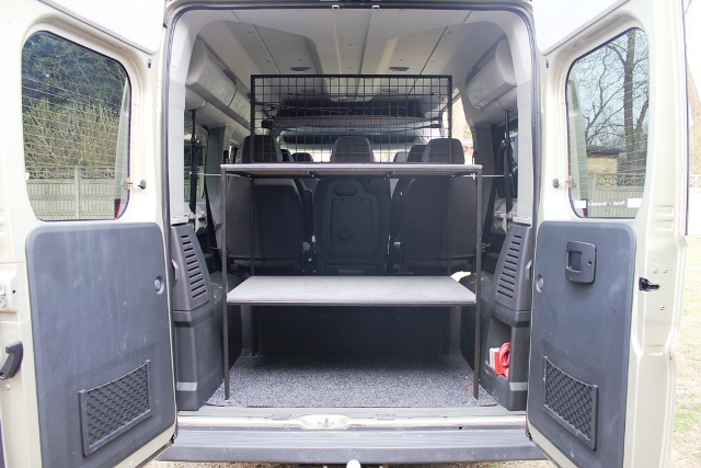 Wnętrze busa z otwartymi drzwiami bagażnika, wyposażone w metalową kratę oddzielającą przestrzeń pasażerską od bagażowej oraz dwupoziomowy metalowy stelaż z półkami.
