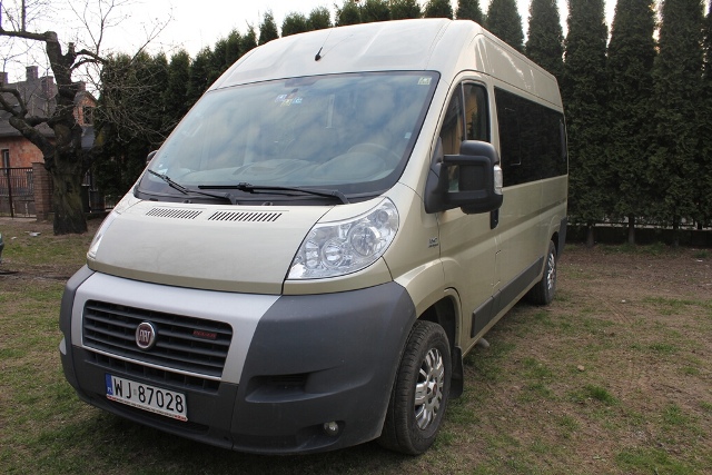 Beżowy bus Fiat Ducato z ciemnymi szybami, zaparkowany na trawiastym terenie, z widoczną tablicą rejestracyjną.