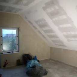 Poddasze w trakcie prac wykończeniowych: widoczne płyty kartonowo-gipsowe na skośnym suficie, ściany przygotowane pod malowanie, okno z widokiem na osiedle, worki z odpadami budowlanymi i odkurzacz...