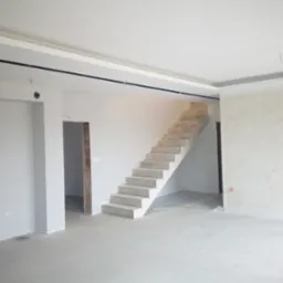 Surowe wnętrze w trakcie wykańczania: betonowe schody bez balustrady, ściany przygotowane do malowania z widocznymi przewodami elektrycznymi i gładzią, sufit z czarnymi elementami oświetleniowymi.