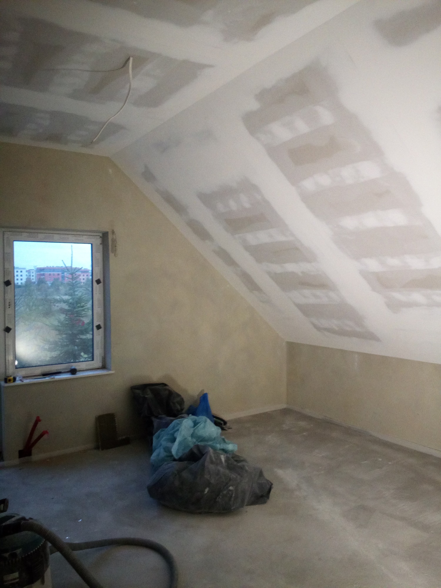 Poddasze w trakcie prac wykończeniowych: widoczne płyty kartonowo-gipsowe na skośnym suficie, ściany przygotowane pod malowanie, okno z widokiem na osiedle, worki z odpadami budowlanymi i odkurzacz...