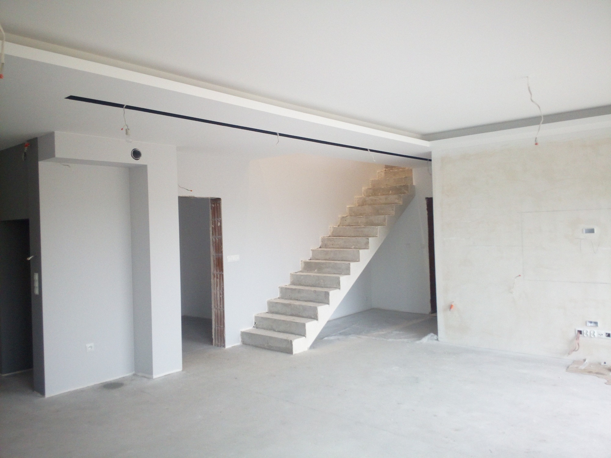 Surowe wnętrze w trakcie wykańczania: betonowe schody bez balustrady, ściany przygotowane do malowania z widocznymi przewodami elektrycznymi i gładzią, sufit z czarnymi elementami oświetleniowymi.