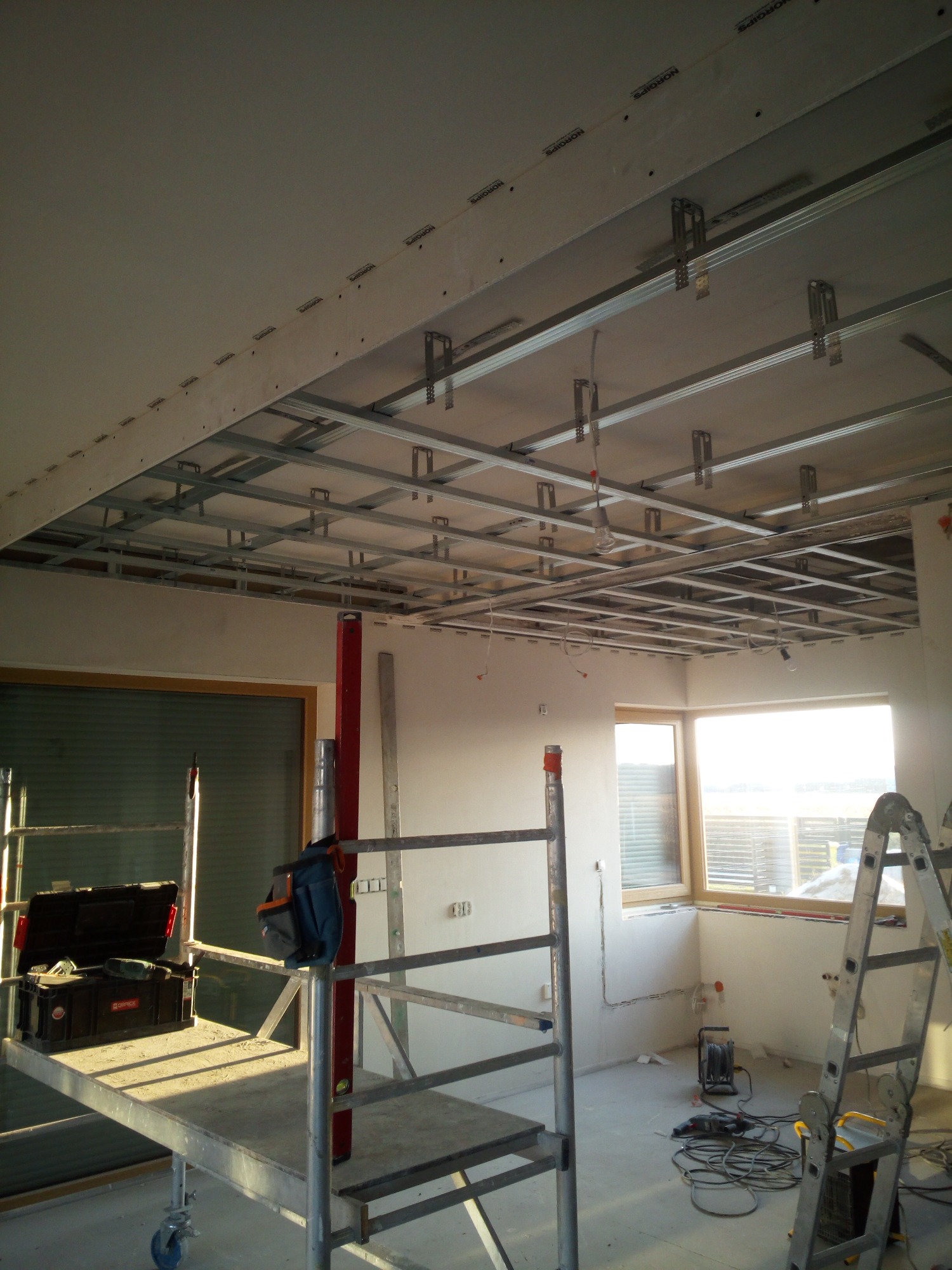 Montaż sufitu podwieszanego z płyt kartonowo-gipsowych na metalowej konstrukcji w jasnym pomieszczeniu, widoczne narzędzia na rusztowaniu i drabina.