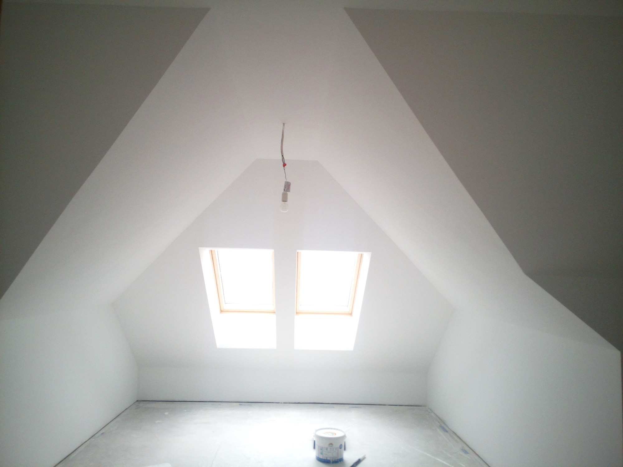Poddasze po malowaniu na biało, z dwoma oknami dachowymi i wiszącą żarówką, na podłodze stoi puszka z farbą.