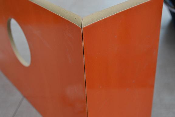 Pomarańczowa płyta z wyciętym okrągłym otworem, połączona pod kątem prostym z drugą płytą, widoczne niedoskonałości powierzchni.