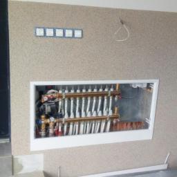 Spm s.c. Michał Malinowski, Paweł Zieliński, Sebastian Kmieć - Rozdzielacz ogrzewania podłogowego w białej wnęce na ścianie o fakturze baranka, widoczne rurki i zawory, z boku czarne drzwi, na ścianie włączniki światła i czujnik dymu.