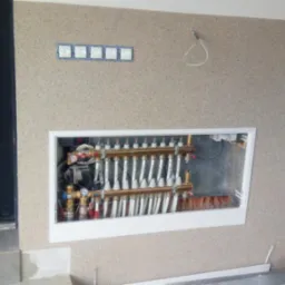 Rozdzielacz ogrzewania podłogowego w białej wnęce na ścianie o fakturze baranka, widoczne rurki i zawory, z boku czarne drzwi, na ścianie włączniki światła i czujnik dymu.