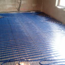 Spm s.c. Michał Malinowski, Paweł Zieliński, Sebastian Kmieć - Niebieskie rury ogrzewania podłogowego ułożone równolegle na folii z czerwoną siatką w surowym pomieszczeniu z oknem i widocznymi przewodami elektrycznymi.