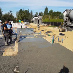 Wylewanie fundamentów domu jednorodzinnego: pracownicy wyrównują świeży beton za pomocą listew wibracyjnych, w tle betoniarka i zabudowa jednorodzinna.