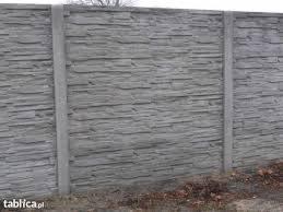 Szary, prefabrykowany betonowy płot imitujący kamień, z widocznymi pionowymi słupkami, na tle ziemi z uschniętymi liśćmi.