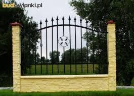 Czarne, metalowe ogrodzenie z dekoracyjnym elementem w kształcie czterolistnej koniczyny, osadzone na niskim, żółtym murku z imitacją kamienia i słupkach z czerwonymi nakryciami.