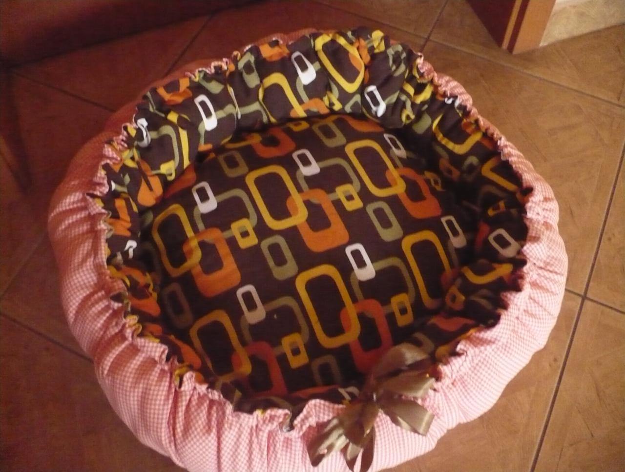 Ręcznie szyte, okrągłe legowisko dla zwierząt z wzorzystą, brązowo-pomarańczową wyściółką i różową obwódką w drobną kratkę, ozdobione kokardką.