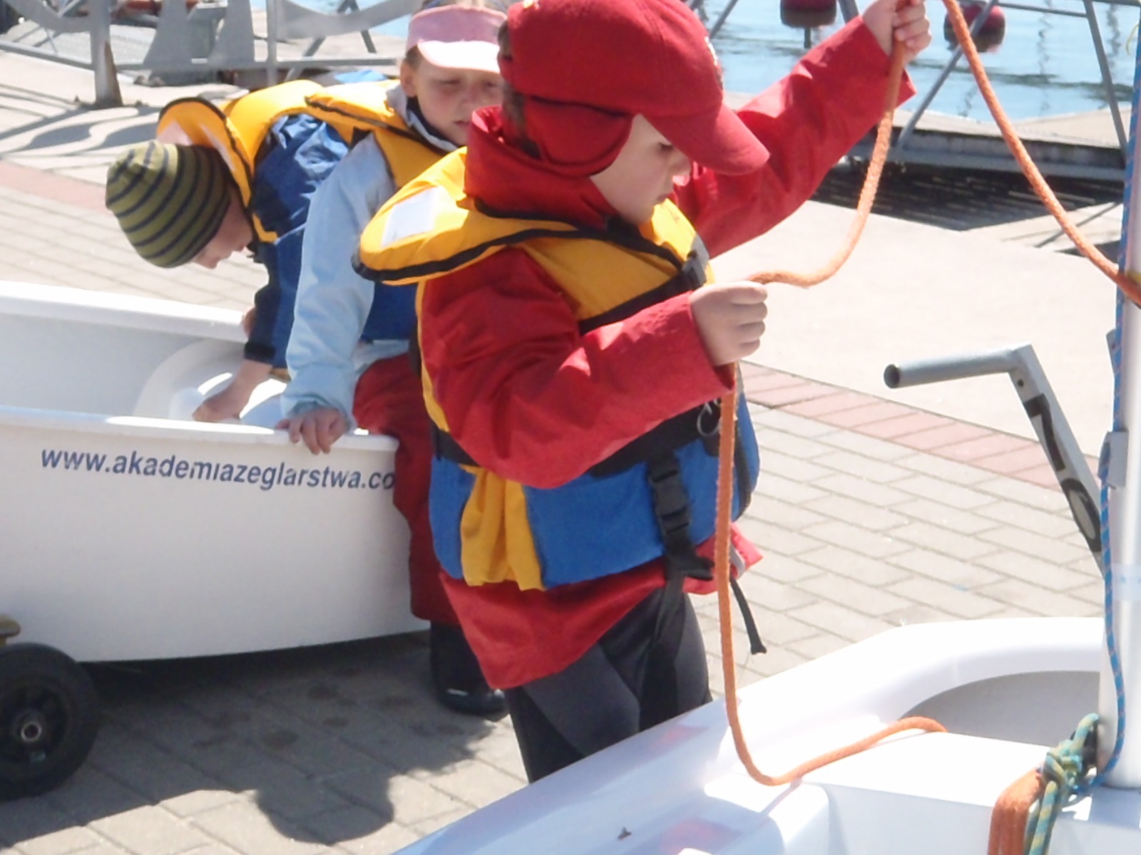 Dzieci w kamizelkach ratunkowych przygotowują się do żeglowania na małych łódkach, widoczny napis 'www.akademiazeglarstwa.co' na burcie łodzi.