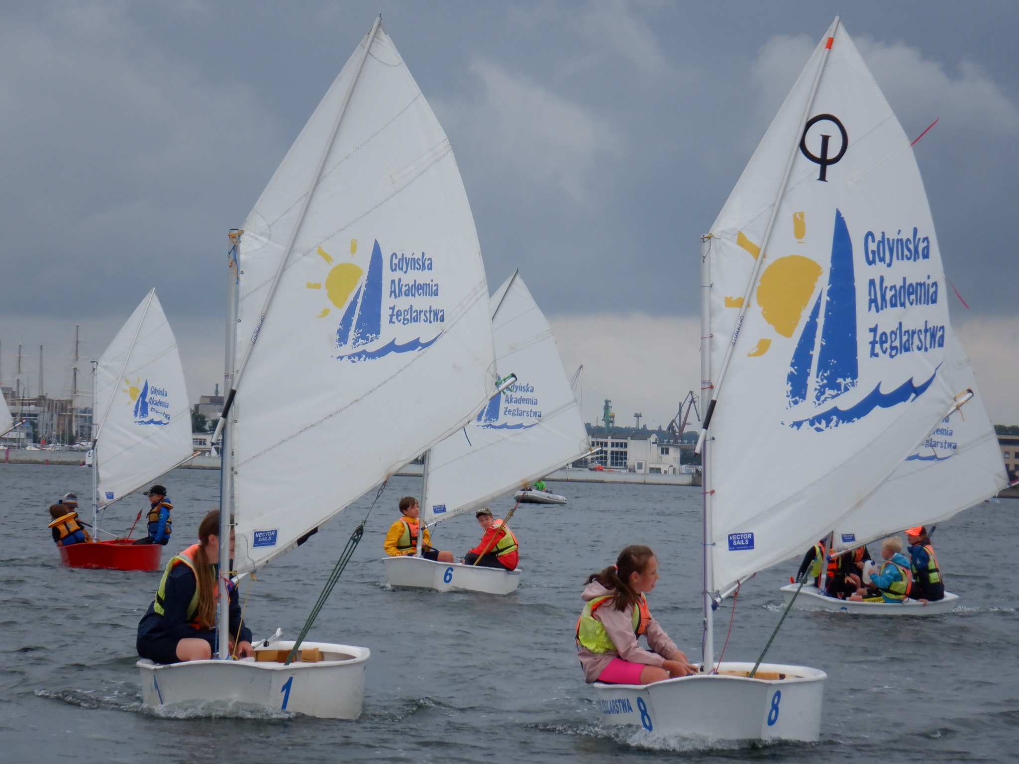 Młodzi żeglarze w małych łódkach żaglowych podczas szkolenia na wodach Zatoki Gdańskiej, żagle z logo Gdynska Akademia Zeglarstwa, pochmurne niebo w tle.