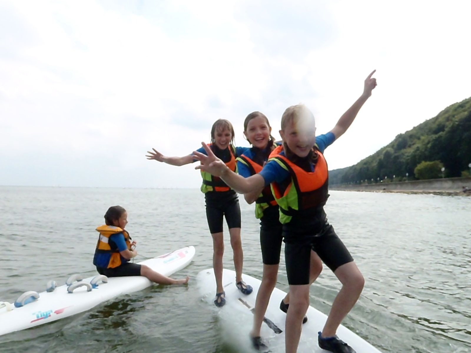 Grupa dzieci w kamizelkach ratunkowych na deskach windsurfingowych, jedna siedzi, trzy stoją z rozłożonymi ramionami, w tle linia brzegowa z drzewami, szary dzień.