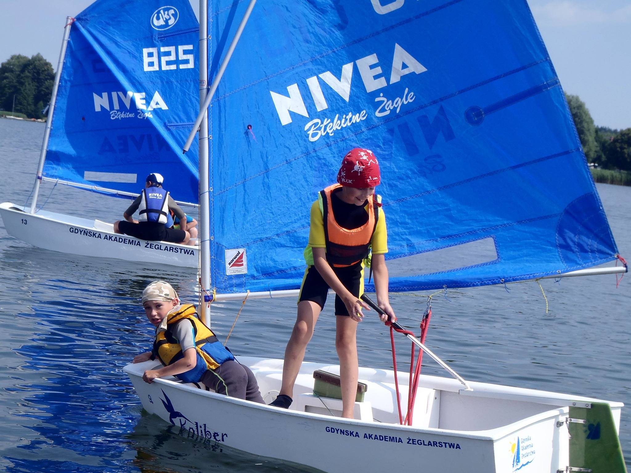 Dwoje dzieci na białych łódkach żaglowych z niebieskimi żaglami, na których widnieje logo Nivea Błękitne Żagle, podczas zajęć żeglarskich na jeziorze, w tle zielone drzewa.