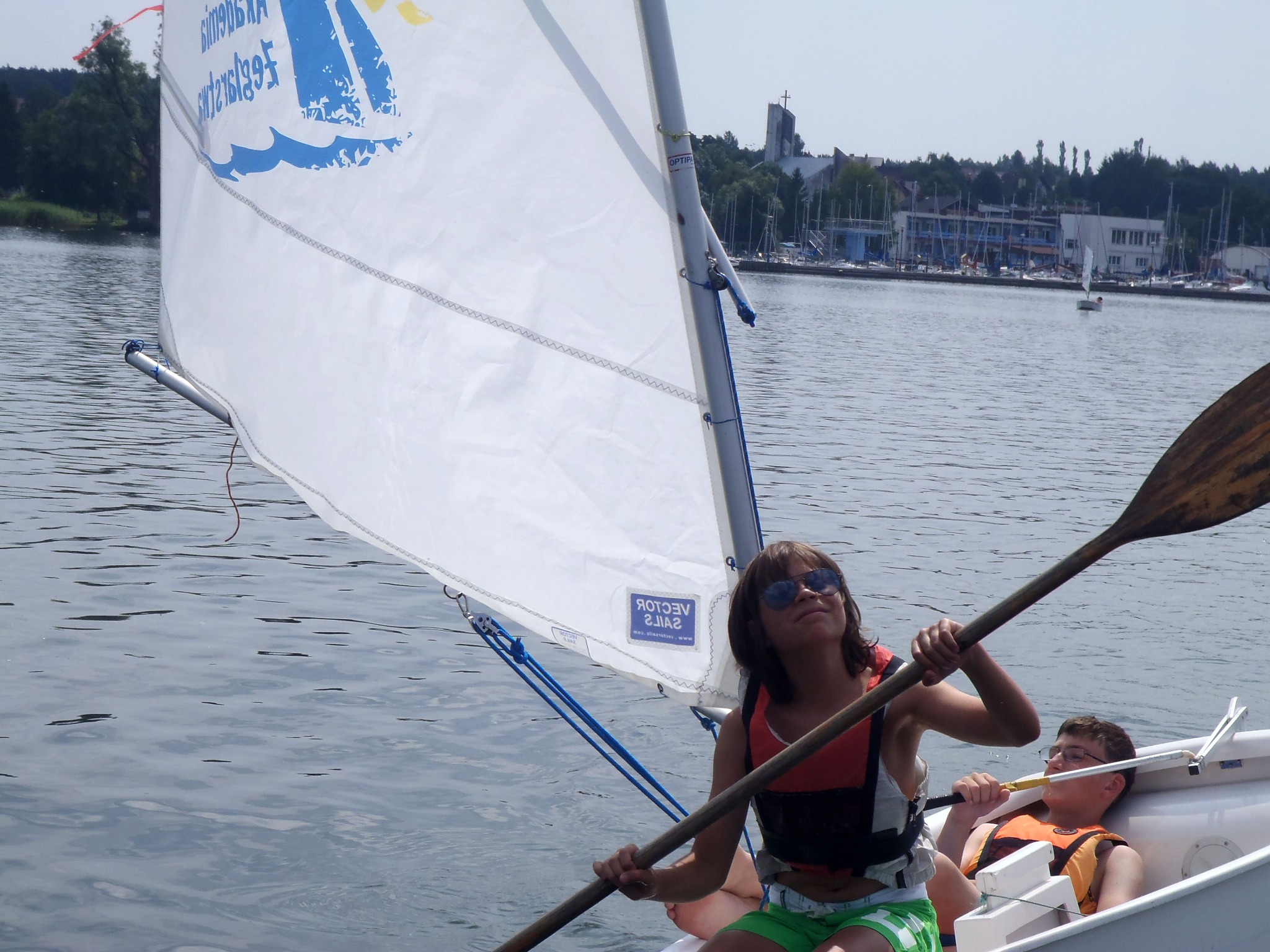 Dziewczyna w okularach przeciwsłonecznych wiosłuje na małej żaglówce z kolegą na jeziorze, w tle budynki i kościół.