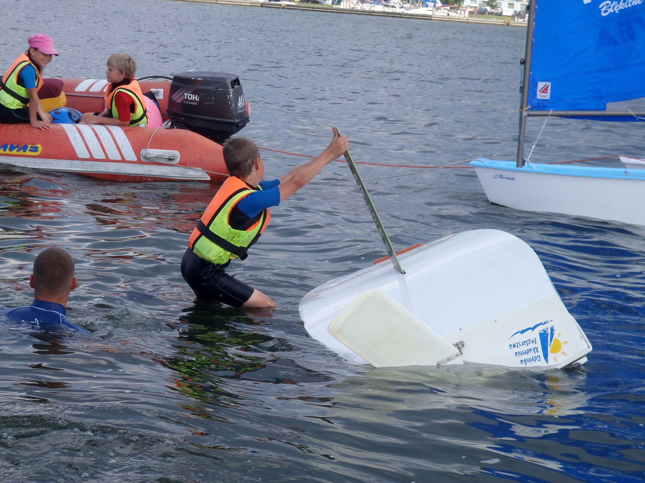 Instruktor pomaga młodemu żeglarzowi odwrócić wywróconą małą żaglówkę na wodzie, w tle ponton z dziećmi w kamizelkach ratunkowych i silnikiem zaburtowym.