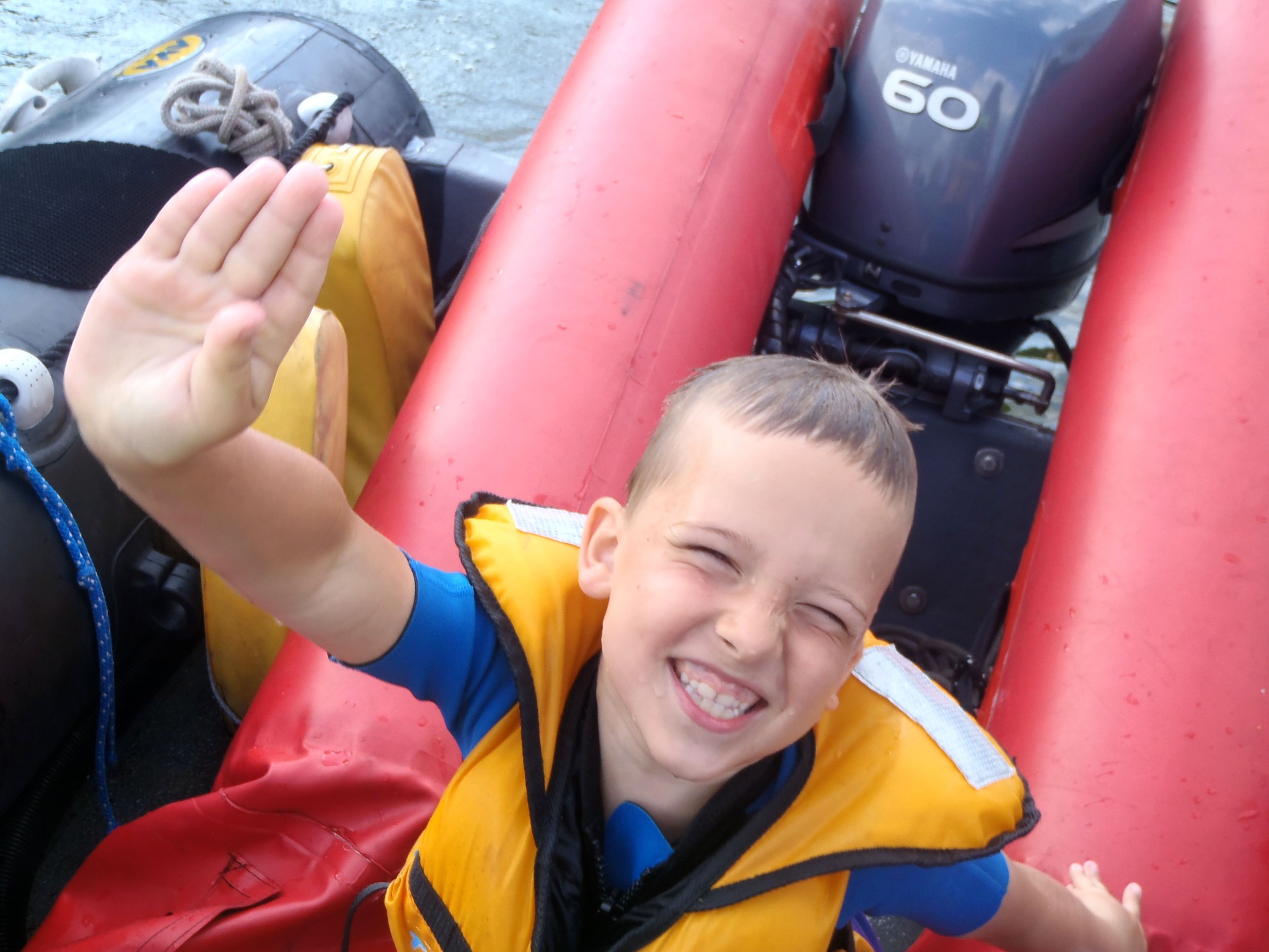 Uśmiechnięty chłopiec w żółtej kamizelce ratunkowej macha ręką, siedząc w czerwonej łodzi pontonowej z silnikiem Yamaha 60.