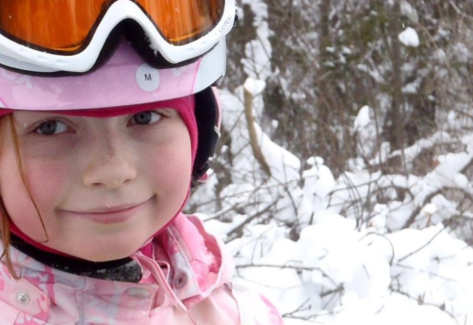Portret uśmiechniętej dziewczynki w różowym kasku narciarskim z goglami na tle ośnieżonych krzewów.