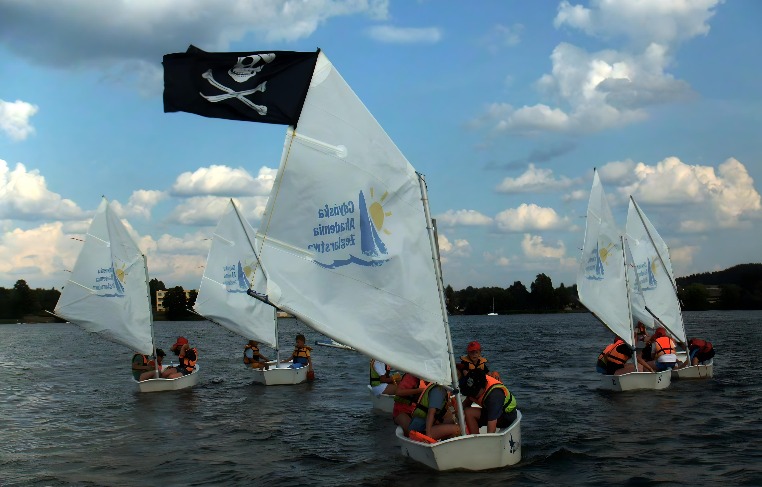 Grupa dzieci w pomarańczowych kamizelkach asekuracyjnych żegluje na małych łódkach po jeziorze, jedna z łódek ma piracką flagę z czaszką i skrzyżowanymi piszczelami.
