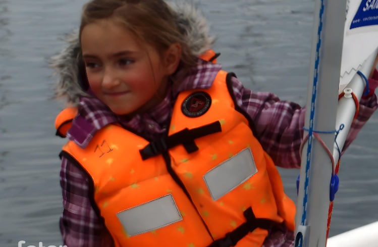 Uśmiechnięta dziewczynka w pomarańczowej kamizelce ratunkowej z numerem '11' i futrzanym kołnierzem, stojąca obok masztu żaglówki na tle wody.